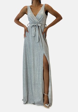 Femme portant une robe portefeuille argentée scintillante sans manches, avec une fente latérale, une ceinture nouée à la taille et des chaussures à talons hauts argentées avec des brides à la cheville.