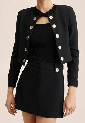 Femme portant un blazer court noir à boutons dorés sur un haut en maille côtelée noir et une jupe noire taille haute avec détails à boutons.