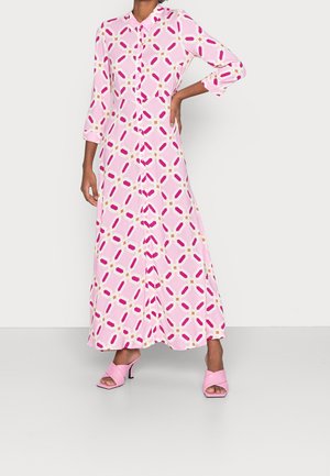 Frau trägt ein langes, rosa Kleid mit geometrischem Muster, 3/4-Ärmeln und rosa Sandaletten mit Absatz, steht mit einer Hand in der Hüfte.