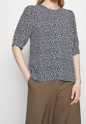 Femme portant un chemisier bleu marine à imprimé floral blanc et manches aux coudes, associée à un pantalon large marron, posant avec une main dans la poche.