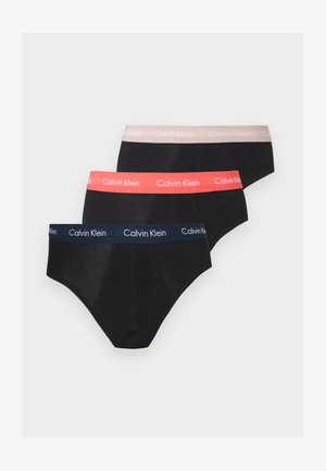 Staand mannelijk model met een donkere huid en kort haar, gekleed in zwarte Calvin Klein slips, poserend tegen een eenvoudige lichte achtergrond.