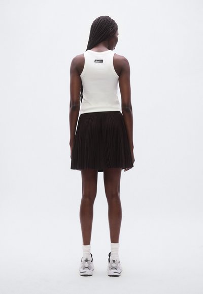 Débardeur blanc, jupe noire plissée, chaussures de sport avec des détails blancs et gris, chaussettes montant à la cheville, vue de dos, fond minimaliste.