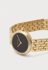 Ρολόι με χρυσό μεταλλικό μπράσελετ, μαύρο στρογγυλό καντράν, χρυσά δείκτες και το λογότυπο "Calvin Klein" στο καντράν.