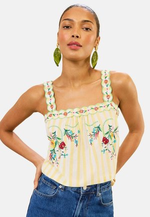 Woman wearing yellow and white striped sleeveless top with floral embroidery and large green leaf earrings, posing with hands on hips.