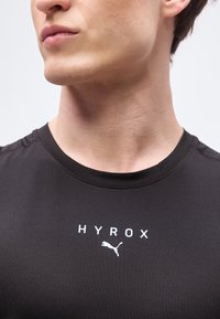 Jeune personne portant un t-shirt noir avec le texte "HYROX" et un logo Puma blanc, montrant le cou et le bas du visage sur un fond uni.