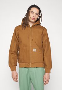 Brown zip-up hoodie made of smooth fabric, featuring a hood, front pockets, and ribbed cuffs. Includes a logo patch on the left pocket.