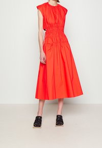 Kvinna klädd i ärmlös, ljusröd midiklänning med samlad midja och svarta öppna mule-sandaler mot en ljusvit bakgrund.