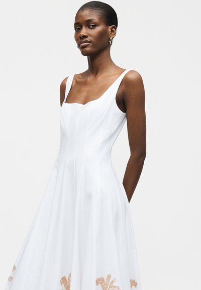 Μαύρη γυναίκα που φοράει λευκό αμάνικο φόρεμα με τετράγωνο λαιμόκοψη και λεπτή καφέ ανθιστή κεντητική κοντά στο τελείωμα, στέκεται μπροστά από απλό φόντο.