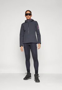 Veste softshell noire avec accents roses, fermeture éclair à l'avant et capuche, associée à un legging noir avec des panneaux texturés et des chaussures de randonnée noires.