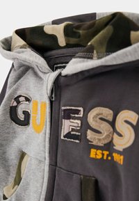 Bluza z kapturem w odcieniach szarości i ciemnych kolorów, z kamuflażowymi akcentami wewnątrz kaptura oraz odważnym haftowanym napisem w różnych kolorach.