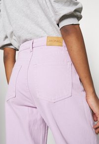 Jeans viola chiaro con vita alta, dotati di tasche posteriori e un'etichetta MONK1 in pelle color beige. Il tessuto ha una superficie liscia e opaca.