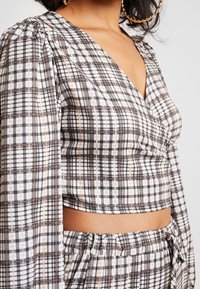Kariertes Cropped-Top mit tiefem V-Ausschnitt, Puffärmeln und tailliertem Schnitt. Die Farben umfassen Schwarz, Weiß und Akzente in Orange.