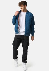 Quilted blå bomberjakke med lynlås, rød foring, ribbede manchetter og krave. Båret over en hvid t-shirt og parret med sorte cargo bukser og hvide sneakers.