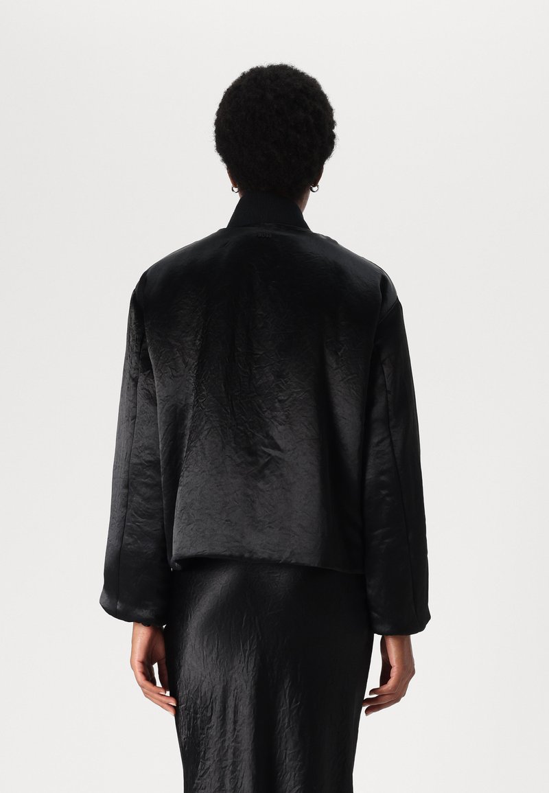 Veste bomber noire en satin avec une texture lisse, épaules tombantes et col côtelé, associée à une jupe noire élégante, vue de derrière.