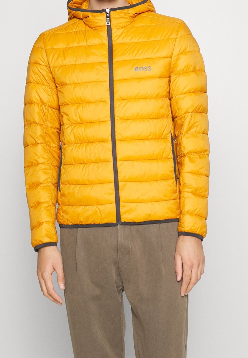 Homme portant une veste matelassée à capuche jaune vif avec des bordures noires et un pantalon gris, debout devant un fond uni.