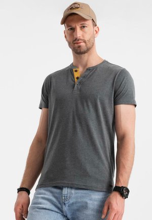 Graues Kurzarm-T-Shirt mit Henley-Ausschnitt, gelben Akzenten und Knöpfen. Getragen mit hellblauen Jeans und einer beigen Mütze.