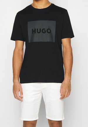 Homme portant un t-shirt noir avec le logo HUGO et un short blanc, debout devant un fond clair uni, les bras le long du corps.