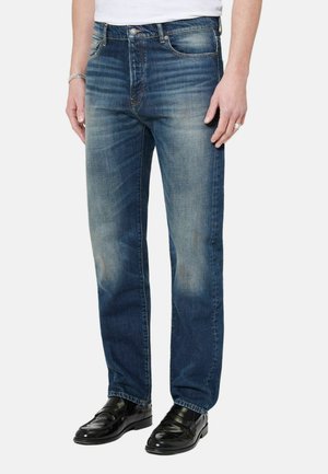 Hombre con jeans azules desgastados, mocasines de cuero negro, camisa blanca, brazalete plateado y un anillo en la mano izquierda.