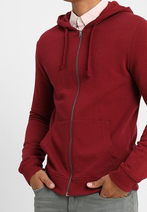 Homme portant un hoodie rouge zippé avec des poches devant, par-dessus une chemise blanche à col, les mains partiellement dans les poches, pantalon gris.