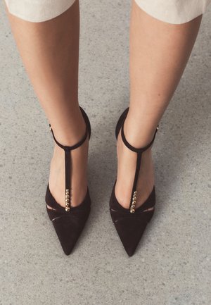 Feet wearing black pointed-toe T-strap heels with gold bead details on a grey speckled floor.