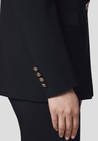 Czarna dopasowana marynarka o gładkiej fakturze, z złotymi guzikami przy mankietach. Zbliżenie na detal rękawa i dłoń.