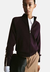 Donna che indossa una giacca con cerniera viola scuro sopra una camicia bianca con colletto, tiene una borsa nera con trama, vestita con pantaloni beige.