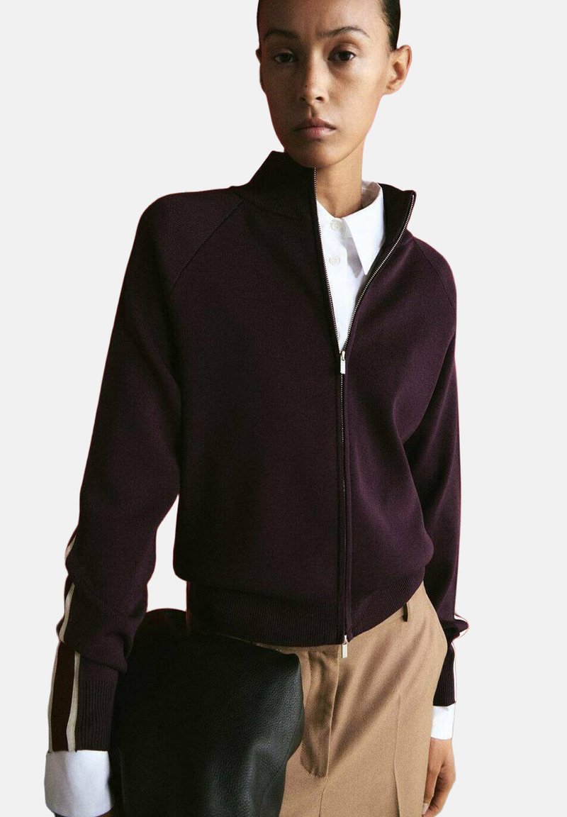 Donna che indossa una giacca con cerniera viola scuro sopra una camicia bianca con colletto, tiene una borsa nera con trama, vestita con pantaloni beige.