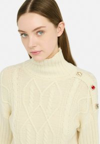 Maglione a collo alto in crema con design a treccia caratterizzato da diverse trama. Accentuato da bottoni colorati simili a gemme su una spalla.