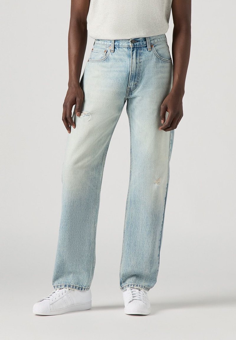 Personne portant un jean bleu clair délavé, droit, avec de légères usures et des baskets blanches sur un fond uni.