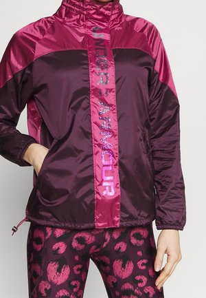 Person iført en mørkelilla og pink Under Armour-jakke med matchende lilla og pink leopardmønstrede leggings, hånd i jakkelommen.