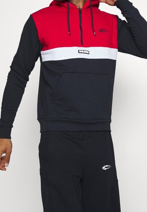 Hombre con una sudadera con capucha roja, blanca y azul marino con un bolsillo delantero y pantalones negros, de pie con un brazo doblado hacia arriba.