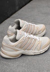 Paar beige und weiße Sportsneaker mit Mesh-Obermaterial und mehreren Streifen, die auf grauem Pflaster liegen.