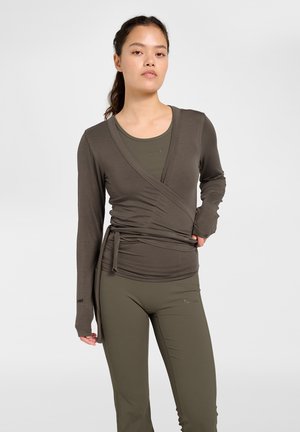 Vrouw draagt een olijfgroene wikkelstijl top met lange mouwen over bijpassende aansluitende legging, staand tegen een effen witte achtergrond.