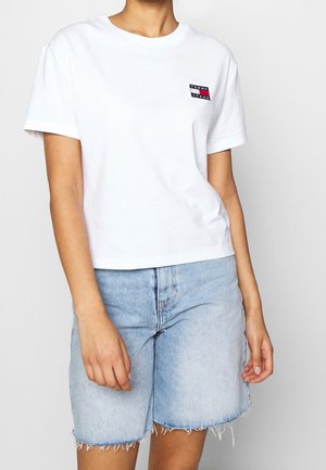 Person iført en hvid Tommy Jeans t-shirt og lyseblå denimshorts med frynset kant, stående mod en ensfarvet baggrund.
