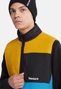 Fleece jakke med gule, svarte og blå seksjoner; har høy krage, glidelåslomme og Timberland-logo på brystet.