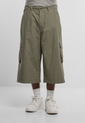 Mand iført olivengrønne vidde cargo shorts kombineret med hvide crew sokker og hvide sneakers på en ensfarvet baggrund.