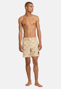 Beige swim shorts with a white geometric pattern, drawstring waist, and a colorful circular patch on the left side. Smooth texture.