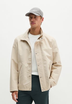 Man met een beige jas, wit overhemd, donkere broek en een zwart-wit geruit petje met handen in de zakken tegen een effen achtergrond.