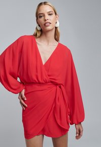Rode wikkeljurk met lange, volumineuze mouwen, een diepe V-hals en een geknoopte taille-detail. Glad, lichtgewicht stof.