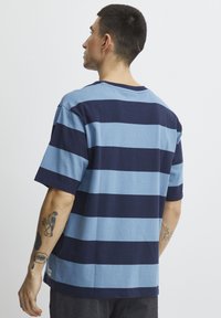 Trumpas rankovėmis T-shirt su mėlynomis ir tamsiai mėlynomis horizontaliomis juostelėmis, pagamintas iš medvilnės, laisvas pritaikymas, su raišteliu apykakle, matosi tatuiruotės ant rankos.