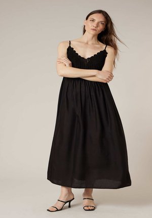 Femme debout, les bras croisés, portant une robe longue noire sans manches avec un corsage en crochet et des sandales à talons noires.