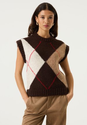 Vrouw met lang bruin haar draagt een mouwloos bruin argyle trui-vest en beige broek, staat met haar handen in de zakken tegen een effen achtergrond.