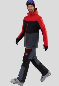 Rød og sort skijakke med lynlåse, grå ski bukser med logo og lynlås-lommer. Modellen bærer skistøvler og skibriller, i en stående stilling.