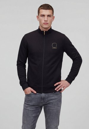 Giovane uomo che indossa una giacca nera con cerniera e un contorno dorato di un volto sul petto, in piedi con una mano in tasca e l'altra sul fianco.