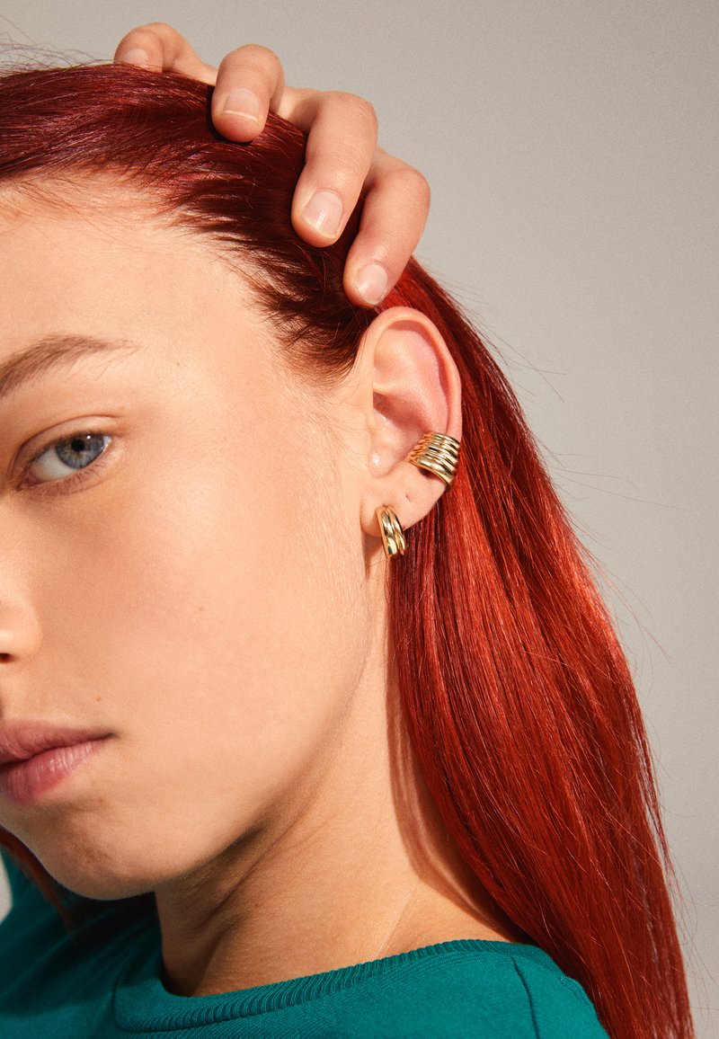 Goldene Creolen mit einem strukturierten, spiralförmigen Design, das eine glatte Oberfläche und einen dezenten Glanz aufweist, getragen am Ohr eines Modells. Rotes Haar ist sichtbar.