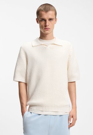 Junger Mann mit kurzen Haaren, der einen strukturierten, cremefarbenen Kurzarm-Strickpullover über einem T-Shirt und hellblauen Hosen vor weißem Hintergrund trägt.