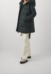 Black puffer coat with a hood, featuring a front zipper, snap buttons, and a green logo detail. Paired with cream-colored pants and gray shoes.