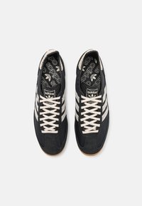 Black suede sneakers with white leather stripes, cream laces, and a black patterned insole, featuring a gum rubber outsole and rounded toe.