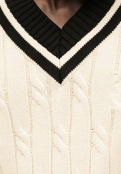 Κοντινή λήψη μιας κρεμ πουλόβερ με πλέξη καλωδίου και λαιμόκοψη V σε μαύρο και άσπρο, φορεμένη πάνω από ένα σκουρόχρωμο, ριγωτό ένδυμα.