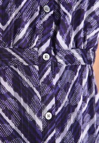 Detailní záběr na vzorovanou látku s diagonálními pruhy v purpurové, bílé a černé barvě, s viditelným zapínáním na knoflíky na levé straně.